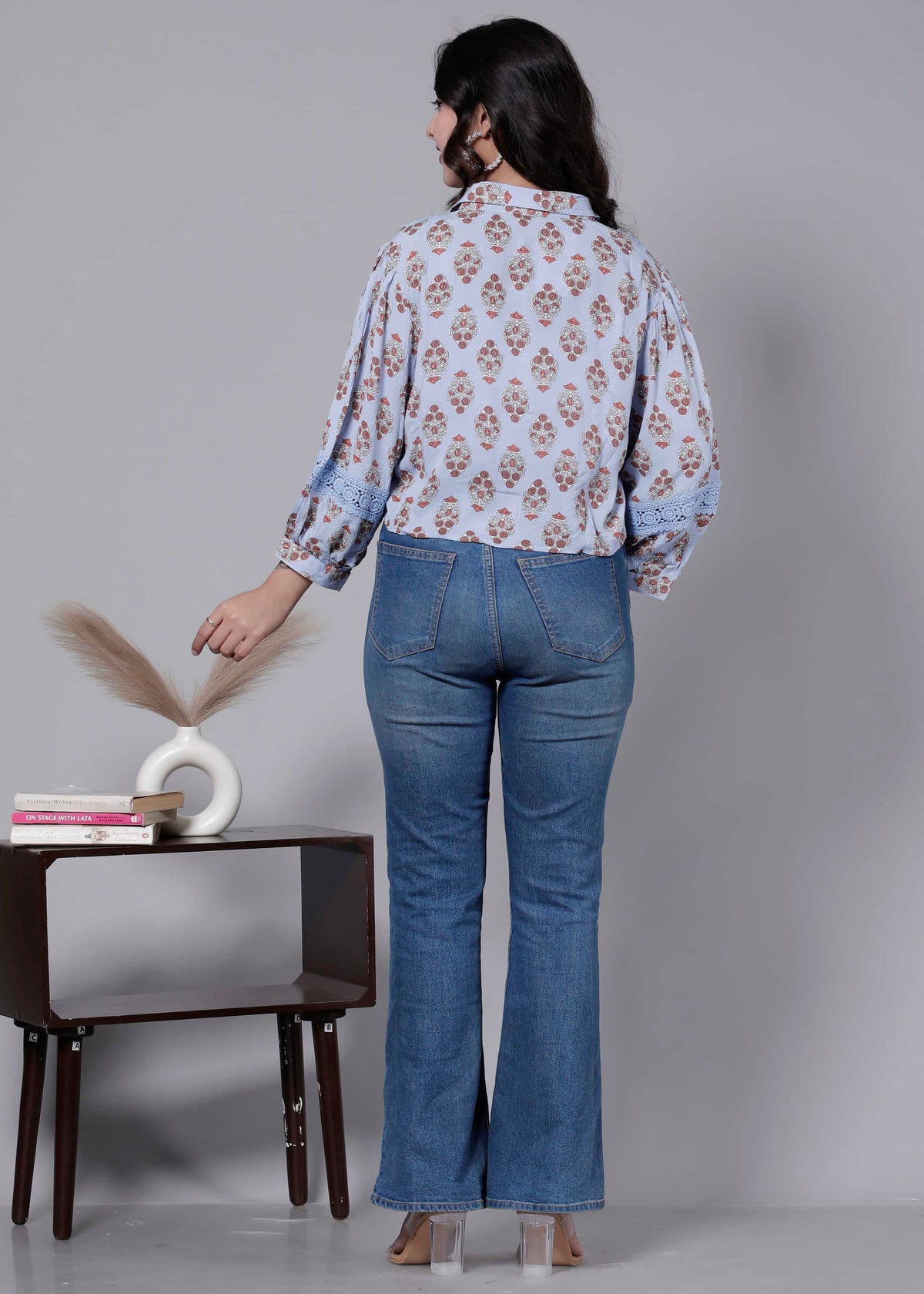 Woman wearing a floral blouse and blue jeans standing in front of a small table with decor items.