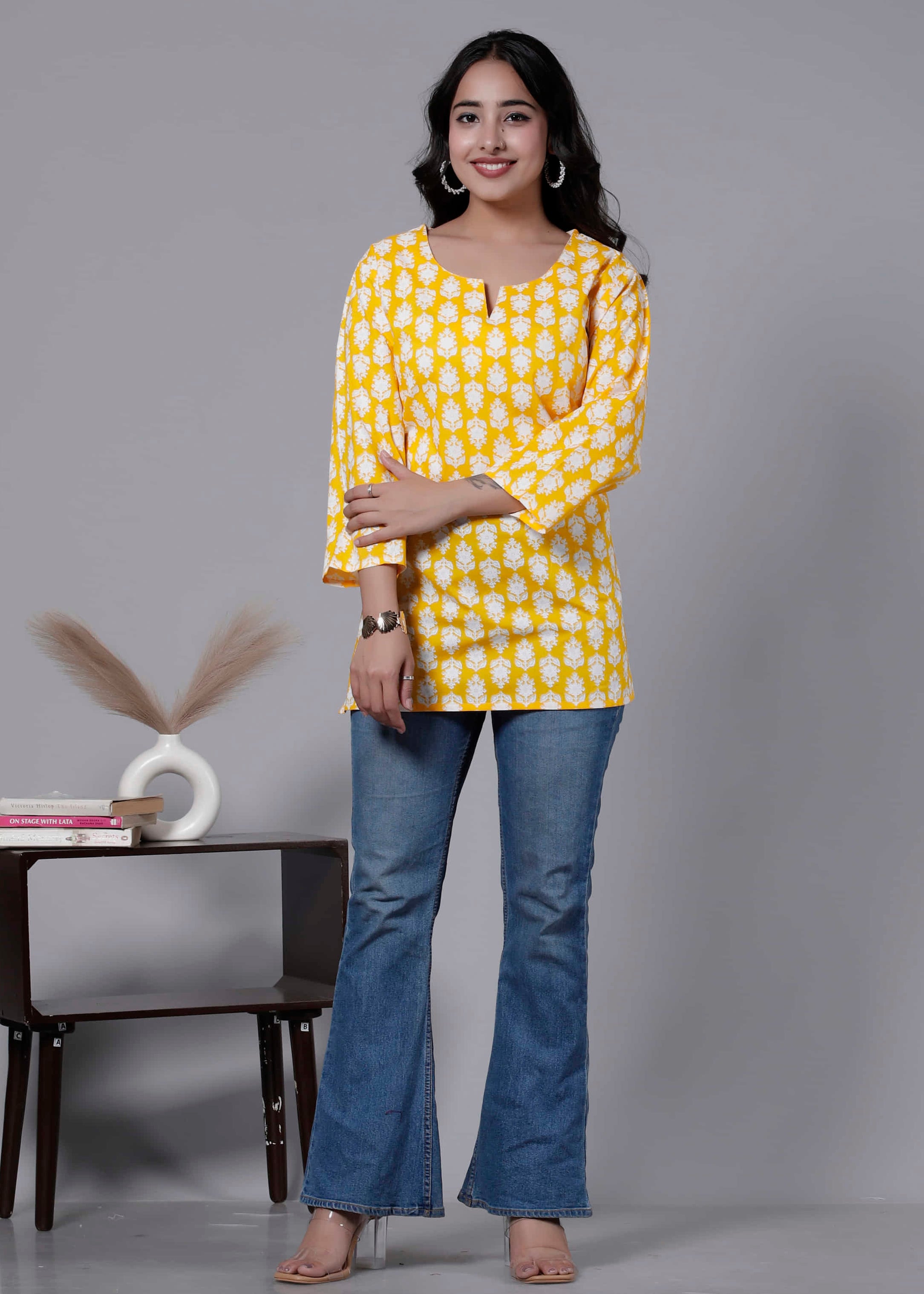 Woman wearing a yellow patterned top and blue jeans standing in front of a gray background with a small table and decorative items.