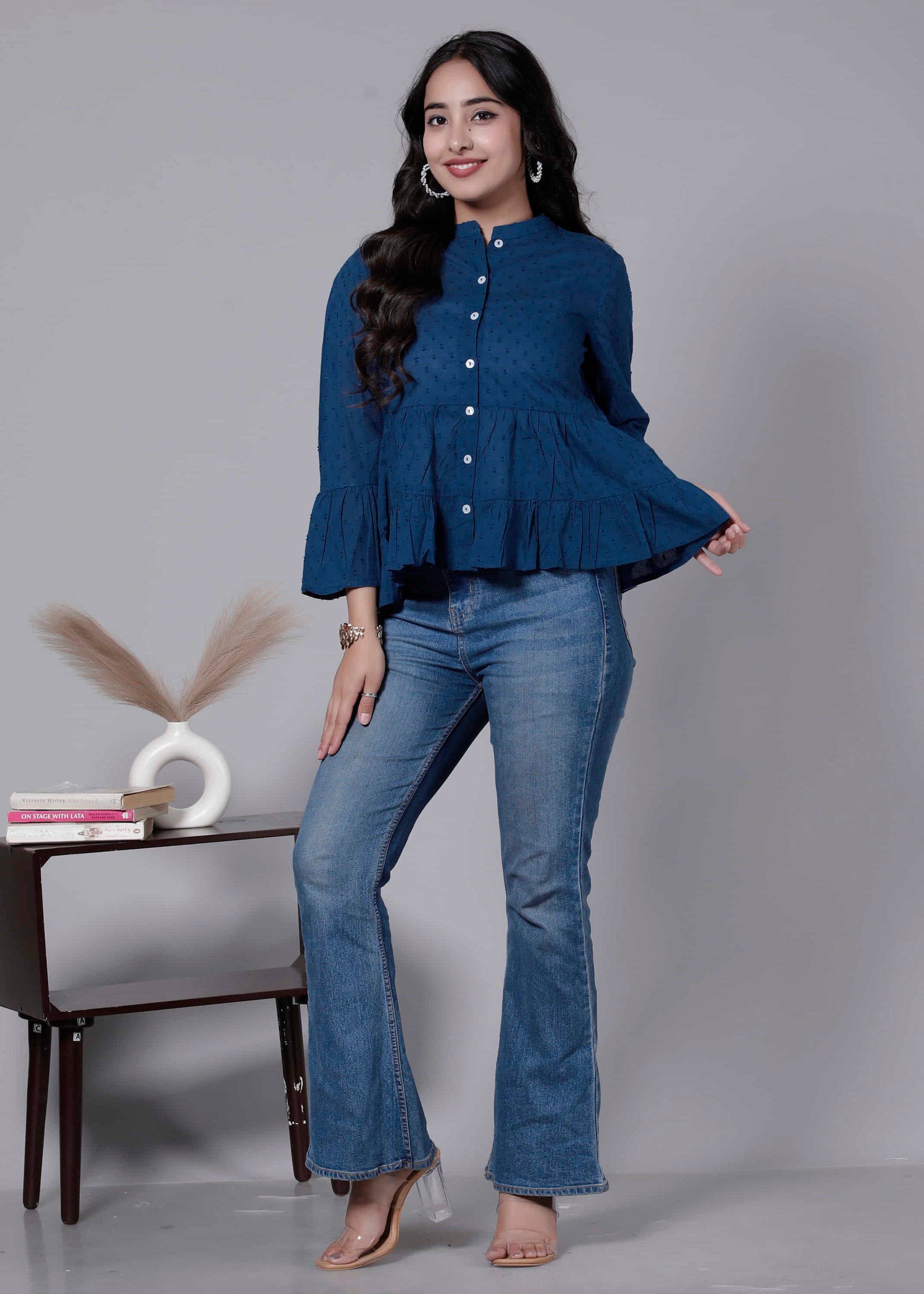 Woman wearing a blue top and jeans standing in front of a gray background with a small table and decorative items.