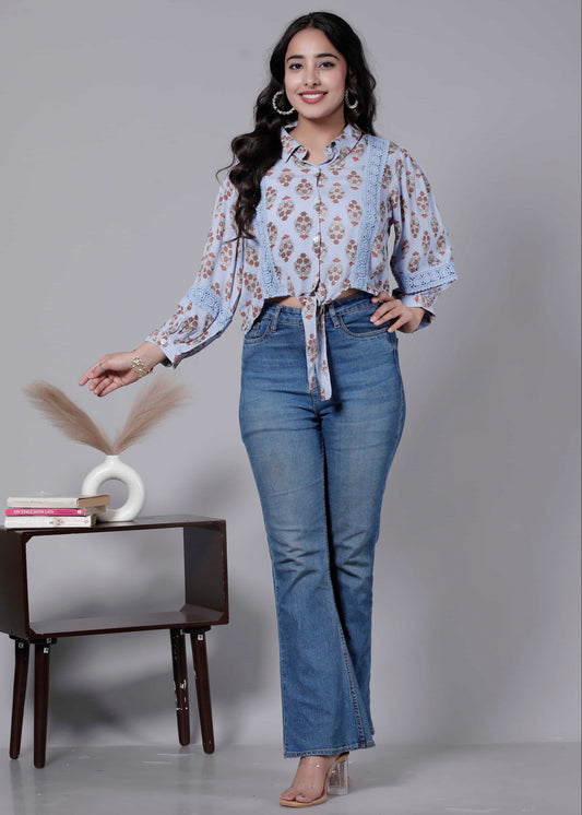 Woman wearing a floral blouse and blue jeans standing next to a small table with decorative items on a gray background